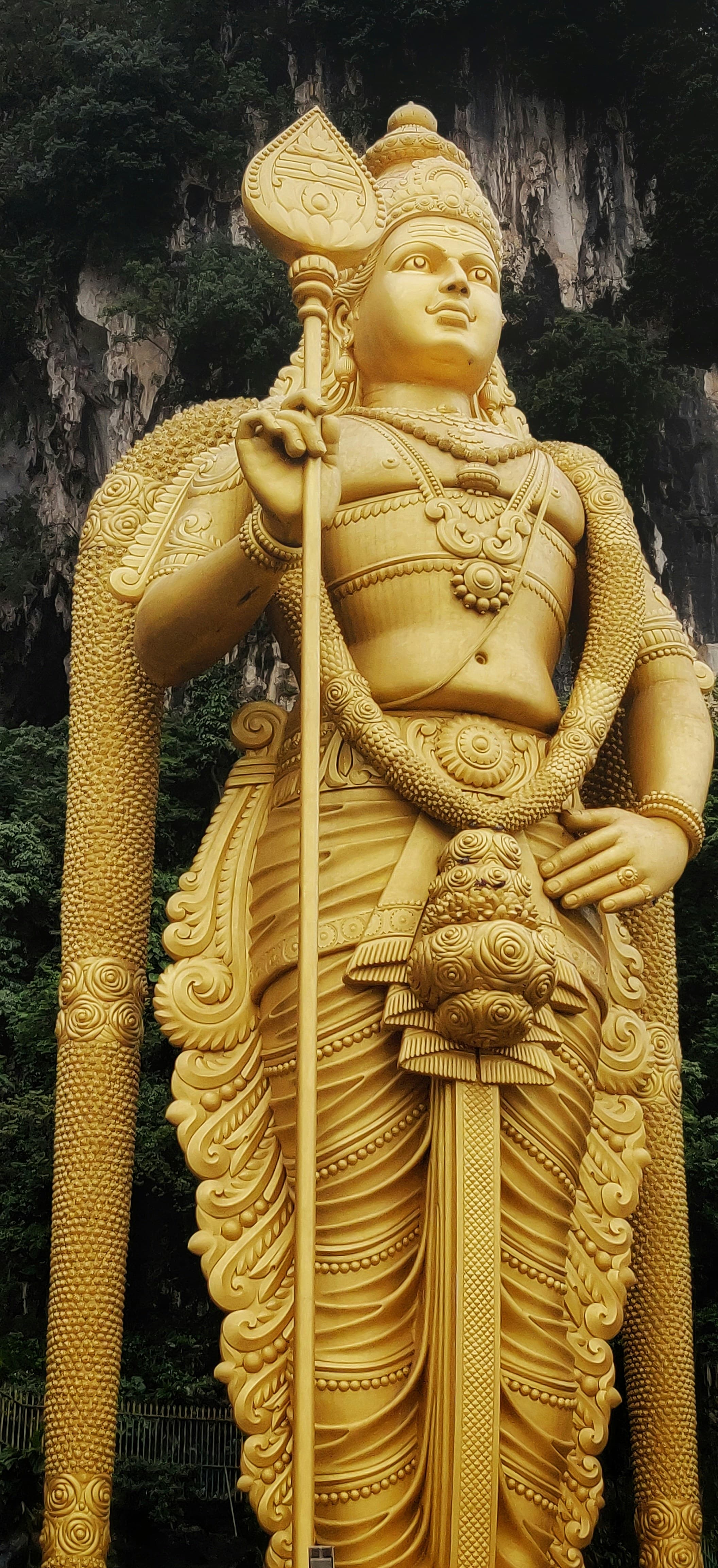 A tall golden statue of Lord Murugan holding a vel, against limestone cliffs