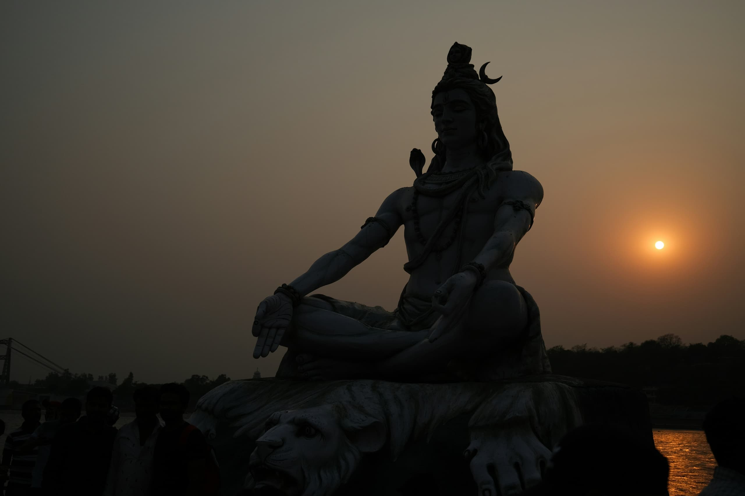 A statue of Lord Shiva seated in meditation on the bank of the Ganga, silhouetted against the setting sun